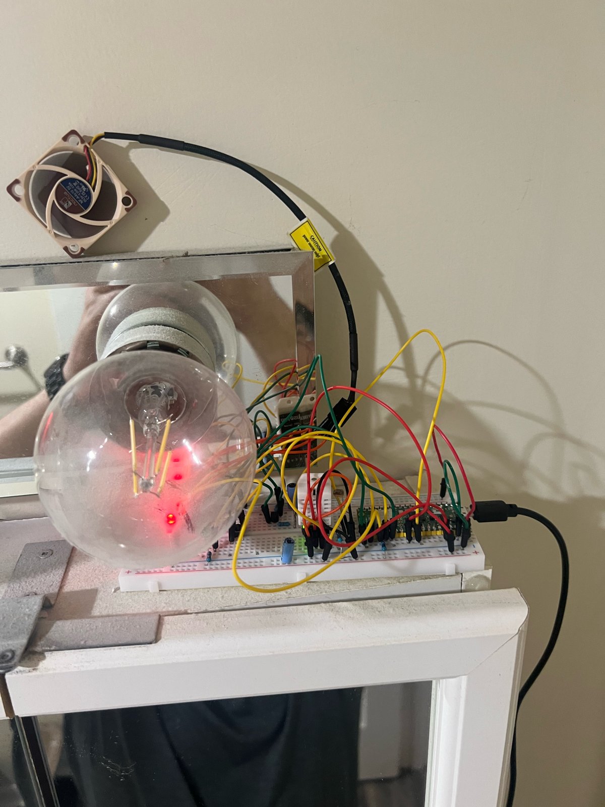 The smart fan build on a bathroom mirror cabinet: a small 40 mm duct fan at top left, a filament light bulb, and a breadboard with a Raspberry Pi Pico 2 W and a tangle of red, yellow and green jumper wires, powered by USB.